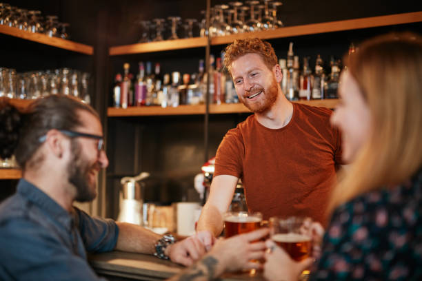 cheerful friends leaning on bar counter, drinking beer and chatting with bartender. night out. - irish culture beer drinking pub imagens e fotografias de stock