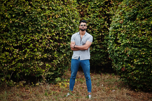 Fashionable tall arab beard man wear on shirt, jeans and sunglasses posed on park against greenery.