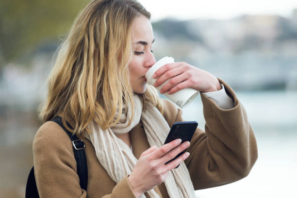istock Pretty young woman drinking coffee while using her mobile phone in the street. 1208629886