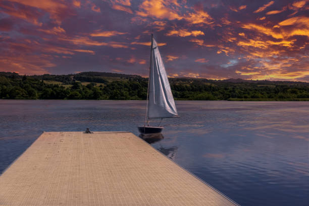 A small sail boat at the side of the flooting pontoon on Loch Semple in Lochwinnoch with the green hills in the background under a blazing red sunset..