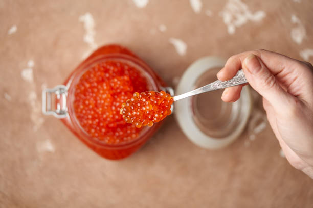 mano femminile con un cucchiaio con caviale rosso su un barattolo di vetro aperto riempito con caviale rosso verso l'alto su una superficie in pietra testurata beige. frutti di mare, prelibatezze, alimentazione sana. posto per il testo. vista dall'alto, pr - vista verso lalto foto e immagini stock