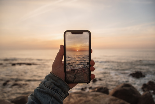 A human hand holding a smartphone to take a picture of a beautiful usnset sky by the sea