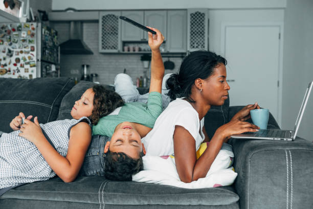 Digital family Mixed race family sitting on the sofa at home and using digital devices. African-american mother using laptop while her two children using smartphones. family dependency mother family with two children stock pictures, royalty-free photos & images