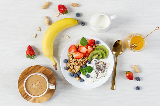 Bowl of granola with yogurt, fresh berries and fruits, cup of coffee on white wooden table background top view. Healthy eating concept.