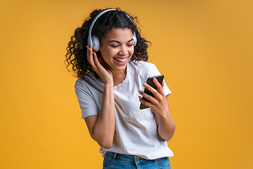 Happy smiling cute dark skinned girl in basic white t-shirt isolated on bright yellow background listening to music using wireless headphones and mobile application on her smartphone. Horizontal shot.