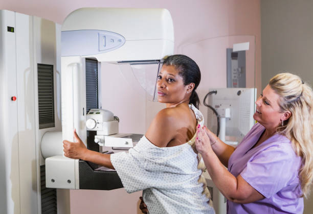 African-American woman getting a mammogram A mature African-American woman in her 40s wearing a hospital gown, getting her annual mammogram.  She is being helped by a technologist, a blond woman wearing scrubs. x ray image medical occupation technician nurse stock pictures, royalty-free photos & images