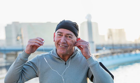 A mature man in his 50s in the city, smiling as he puts on earphones to listen to music.