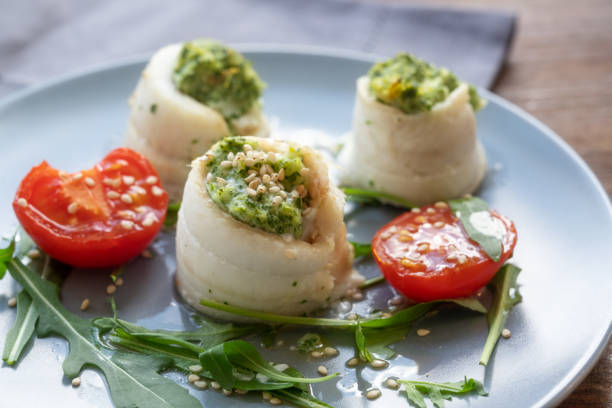 drei fischfiletbrötchen aus scholle gefüllt mit püriertem sellerie und rucola, serviert mit gegrillten tomaten, rucola-salat und sesamsamen auf einem blauen teller, vorspeise für ostern und frühling - flounder fillet seafood meal stock-fotos und bilder