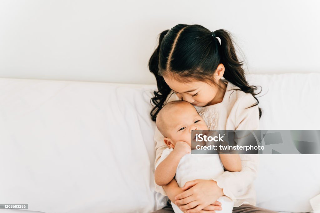 Asian child hugging newborn baby on bed Caring Asian girl in casual wear hugging small brother embracing newborn baby with opened mouth smelling toddler sitting on bed in house Sibling Stock Photo