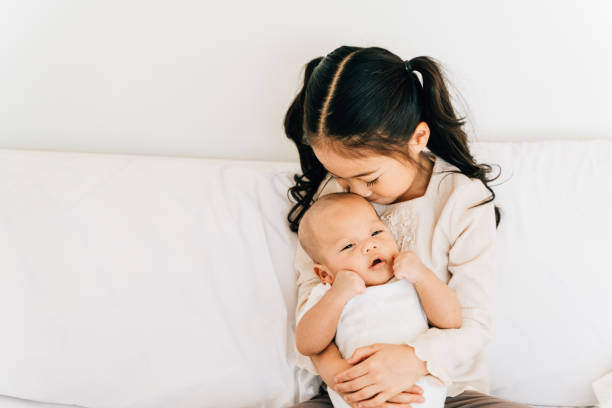 Asian child hugging newborn baby on bed Caring Asian girl in casual wear hugging small brother embracing newborn baby with opened mouth smelling toddler sitting on bed in house sister stock pictures, royalty-free photos & images