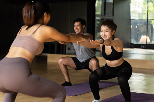 Group of happy Asian women and man doing squat exercises on yoga mats in aerobics class. Young sporty people smiling while working out in gym studio. Fitness Class Concept