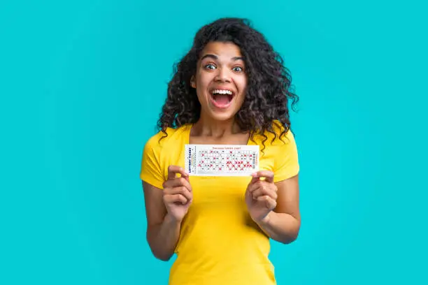 Young cute african american girl wearing bright yellow t-shirt holding lottery ticket in hands looking  excited to get winning results. Studio shot of woman celebrating victory on blue background.