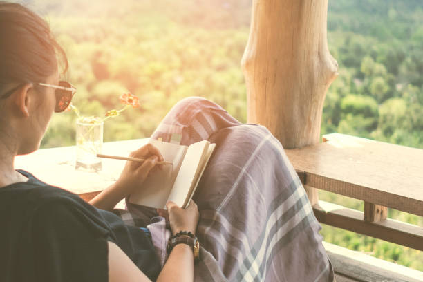 Woman is writing down in small white memo notebook for take a note plan to do or write a book with beautiful terrace of house and nature mountain view background.