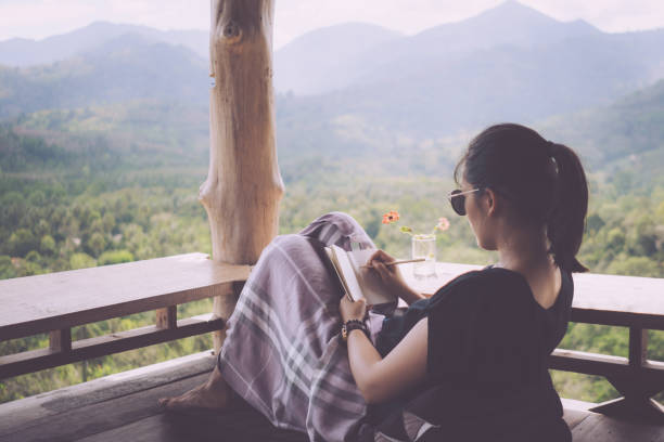 istock Woman is writing down in small white memo notebook for take a note plan to do or write a book with beautiful terrace of house and nature mountain view background. 1208694620