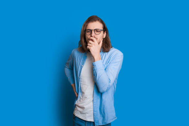 istock Caucasian man with long hair and nice beard is touching his chin while looking aside through his glasses and posing on blue background 1208696854