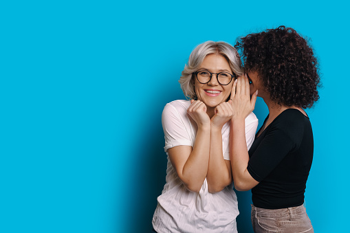 Chica caucásica de pelo rizado está hablando algo secreto a su amiga rubia que está usando anteojos y posando en un fondo de espacio en blanco azul photo