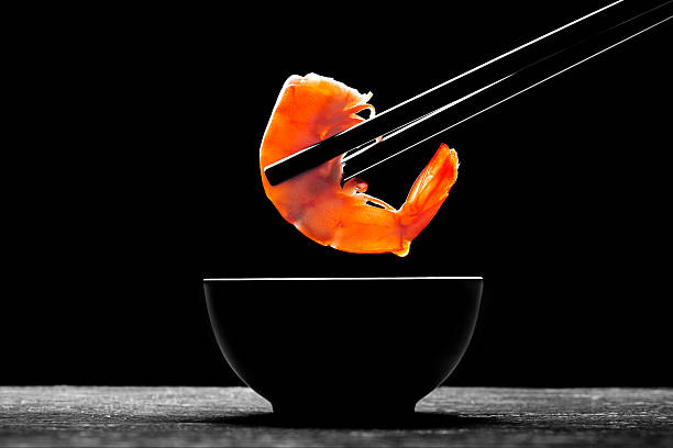 Black chopsticks holding a cooked prawn above a black dish stock photo