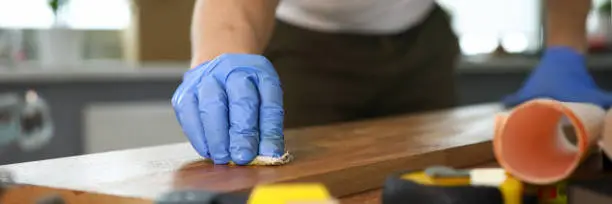 Close-up of male hands in protective gloves grinding wooden surface. Equipment and tools on working table. Carpentry work and joinery processing concept