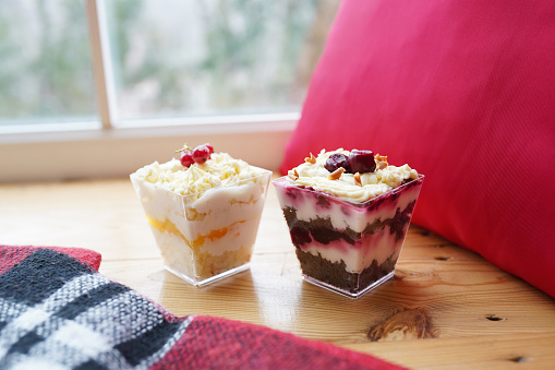 Two colorful biscuit desserts with berries and cream in glasses on table near the window.