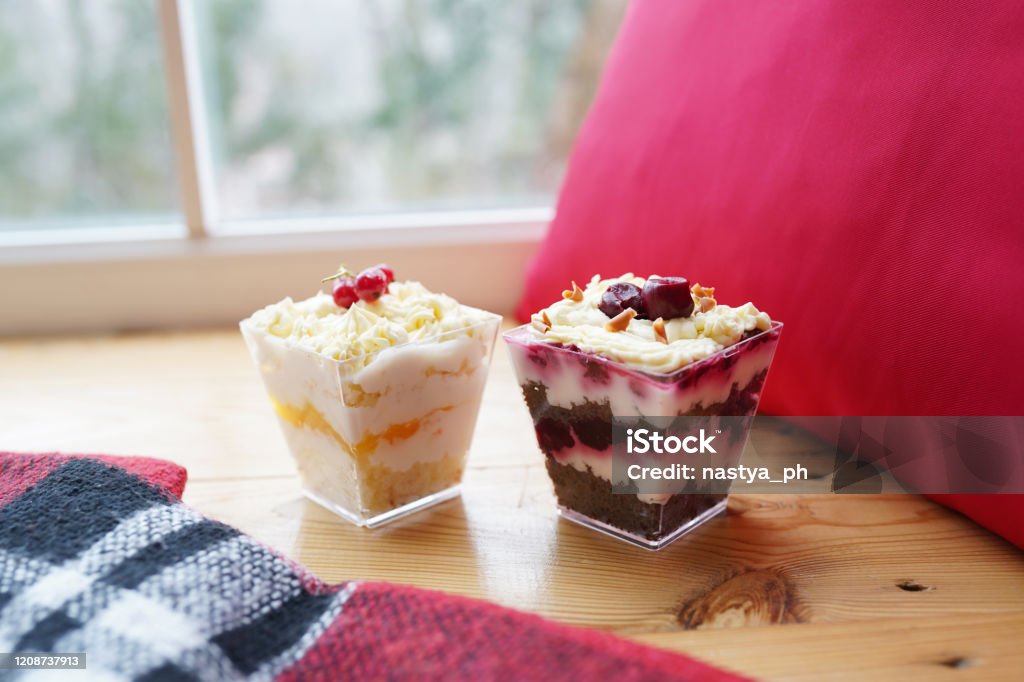 Two colorful biscuit desserts with berries and cream in glasses on table near the window. Cake Stock Photo