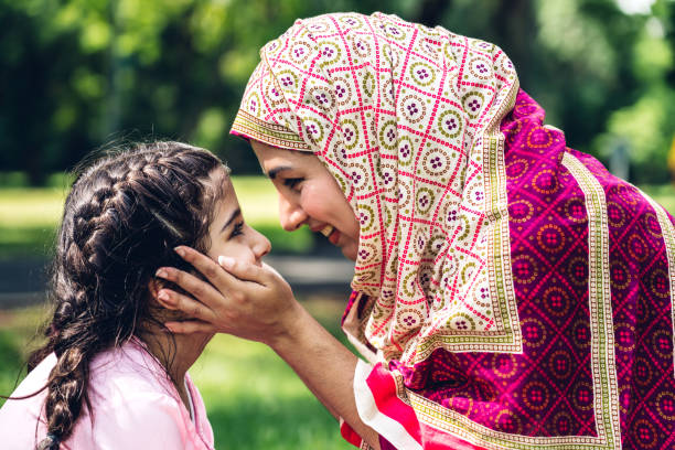 Portrait of happy lovely family arabic muslim mother and little muslim girls child with hijab dress smiling and having fun hugging and kissing together in summer park stock photo