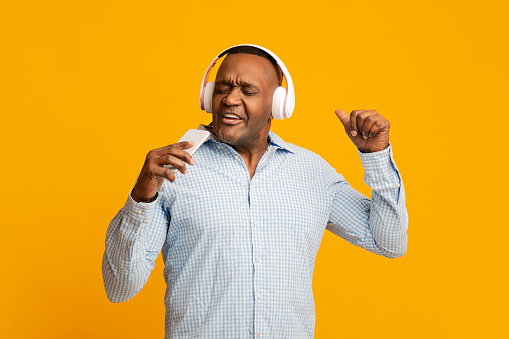 Karaoke lover. Emotional african mature man in headphones singing at phone like a microphone, orange studio background