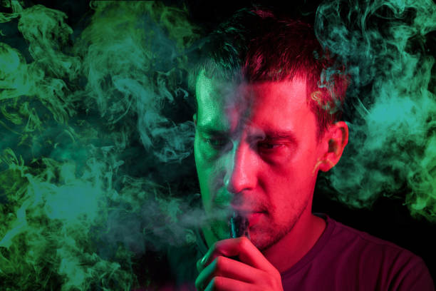 istock A man holds in his hand and drags on an electronic cigarette with a vape while smoking and releases smoke to the sides, his face is highlighted with green neon chemical light, and head is in clouds 1208751903