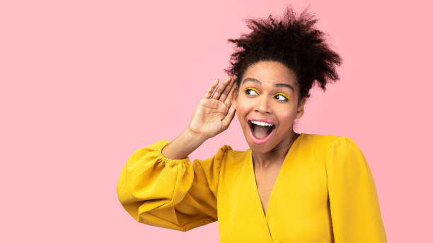 Beautiful afro girl holding hand near ear Gossip Concept. Interested black girl putting hand to her ear to hear better, listening over pink wall, copyspace eavesdropping stock pictures, royalty-free photos & images