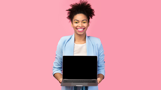 Useful Website. Cheerful afro girl holding laptop computer, showing blank screen over pink studio wall, mock up