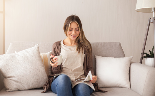 Home and leisure concept. Smiling woman sitting on couch and reading magazine with coffee at home