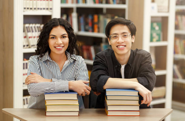 책 더미에 기대어 웃고 있는 두 명의 다문화 학생 - bookstore student chinese ethnicity book 뉴스 사진 이미지