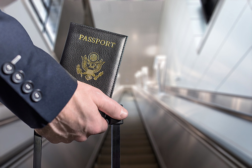 Man (businessman)  in a blue suit with suitcase holding american passport  on the airport escalator