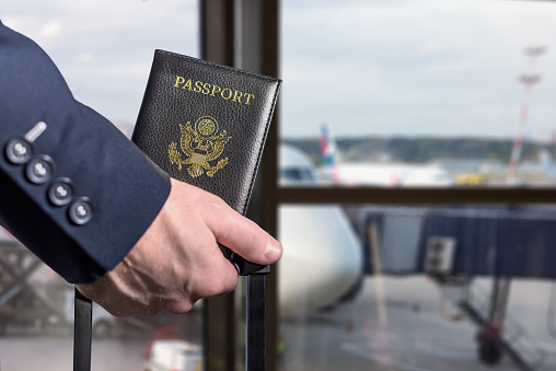 Man (businessman)  in a blue suit with suitcase holding american passport in the airport waiting zone looking through the window at planes.