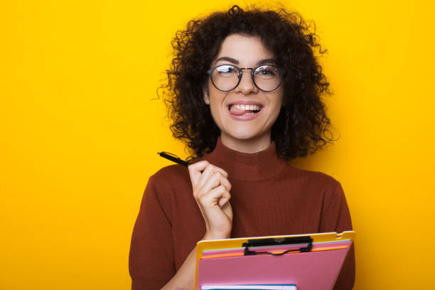 istock Curly haired brunette student looking through eyeglasses is holding a pen and some books while posing on a yellow background 1208760560