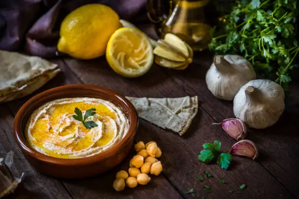 High angle view of a homemade hummus surrounded by the ingredients for cooking and seasoning the dipping sauce like lime, chick pea, garlic, parsley, and olive oil on a rustic wooden table. Low key DSLR photo taken with Canon EOS 6D Mark II and Canon EF 24-105 mm f/4L
