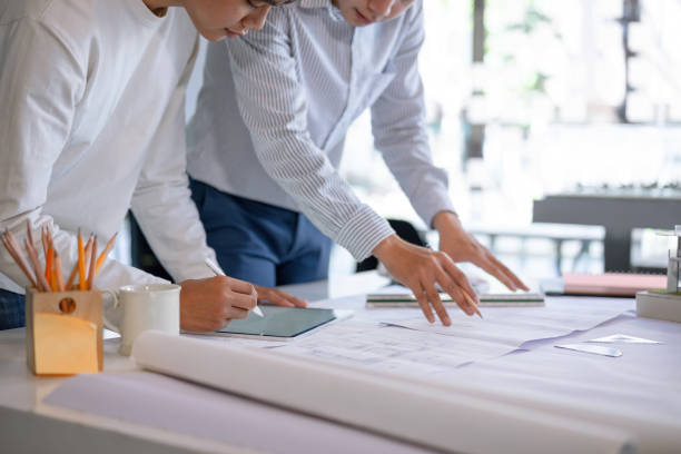 istock Young asian architect colleagues discussing about construction plans on blueprint and tablet at construction site office. Architect using technology concept. 1208761984