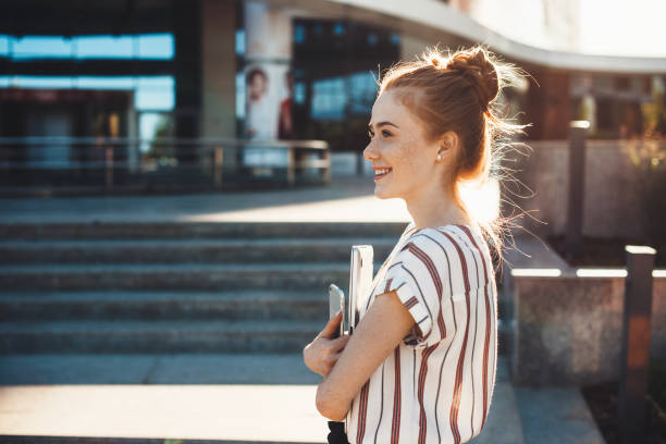 cheerful caucasian girl with red hair and freckles is holding a tablet and looking away staying in front of a building - estudante universitária imagens e fotografias de stock