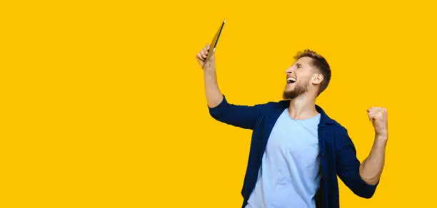 Caucasian man with beard and red hair is looking at his tablet and shouting being happy and posing on a yellow background with freespace