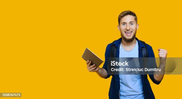 Caucasian Student With Red Hair Nd Nice Beard Is Holding A Tablet And Wearing A School Bag While Gesturing A Win On A Yellow Free Spaced Wall Stock Photo - Download Image Now