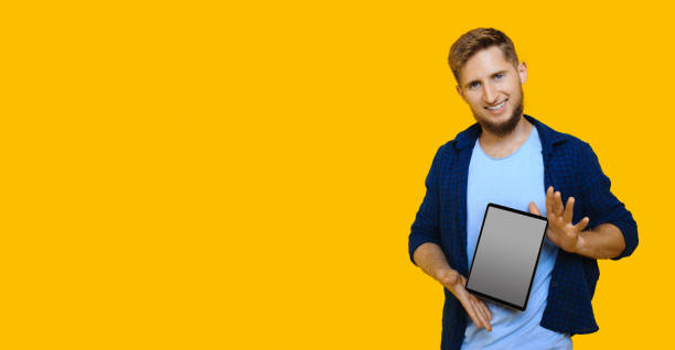 istock Bearded caucasian man with red hair is advertising something using a tablet with blank space on a yellow wall 1208767523
