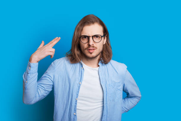 istock Caucasian man with long hair and eyeglasses gesturing with his hand like shooting while posing on a blue background 1208767677