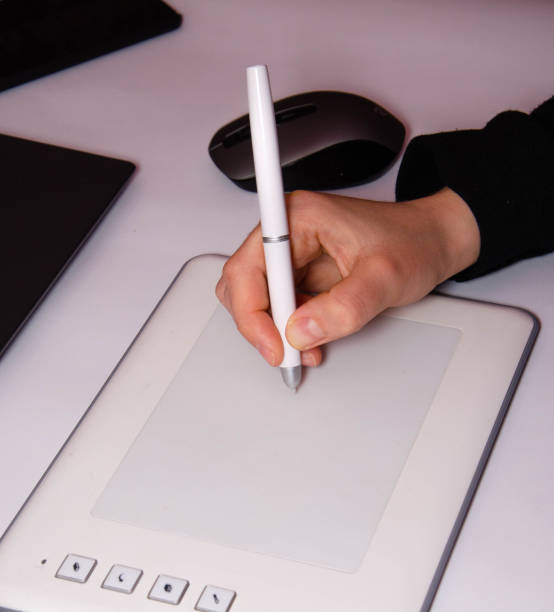 istock Female hands work on a graphic tablet. Hand holds stylus pen and draws. White graphic tablet. The work of a graphic designer. Girl works on a tablet connected to a laptop. Rear view from  behind 1208780269