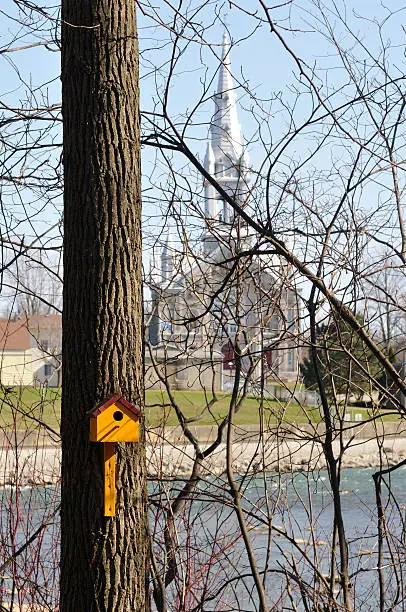 Contrast between a modest yellow birdhouse and, in background, a majestic church, in Chateauguay, Quebec, Canada.