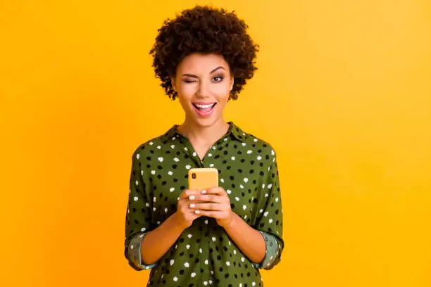 Photo of Portrait of excited crazy afro american girl hold smart phone use read social network news wear stylish green bright clothing isolated over yellow color background