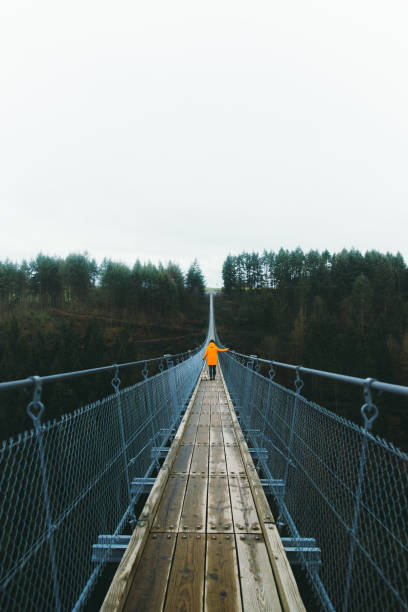 Woman and a dog walking at the suspension bridge enjoying the scenic view of German nature Young woman in orange raincoat and her small cute dog - pug breed hiking on the big and beautiful suspension bridge in Germany man walking a pug dog stock pictures, royalty-free photos & images
