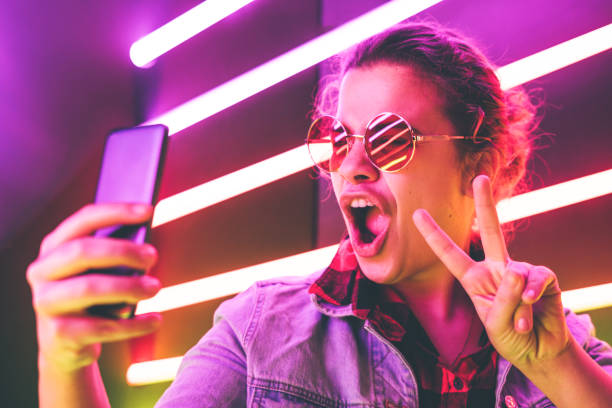 Portrait of modern young woman in glasses making selfie at the neon light. Portrait of modern young girl in round glasses making selfie at the neon light. Shows sign pis. selfie girl stock pictures, royalty-free photos & images