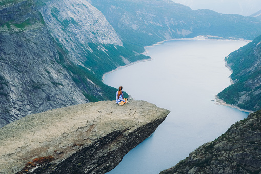 Young woman hiker and her small cute dog - pug breed feeling freedom and happiness sitting on the cliff at the edge of the Trolltunga mountain enjoying the view of dramatic sunrise above the fjord in Norway