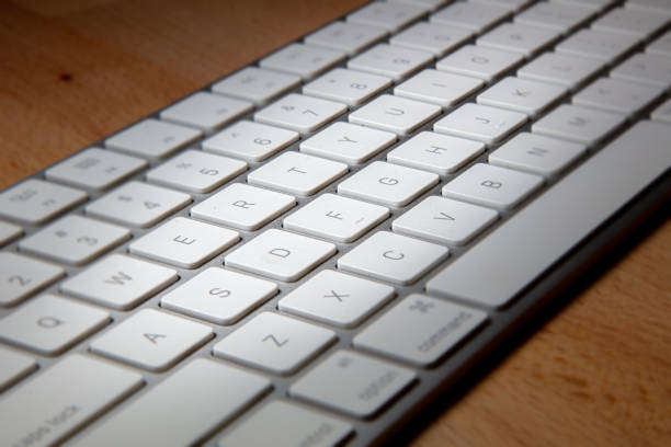 A close up of a computer keyboard on a wooden desk. Macro photography of a modern white aluminium keyboard on wood. Sharp detailed capture. Focused on the key. White keys with grey letters