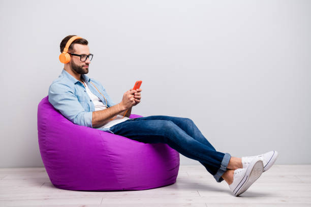Full length profile photo of funny guy sitting comfy armchair holding telephone using cool earflaps choose song wear specs casual denim outfit isolated grey color background Full length profile photo of funny guy sitting comfy armchair holding, telephone using cool earflaps choose song wear specs casual denim outfit isolated grey color background bean bag stock pictures, royalty-free photos & images