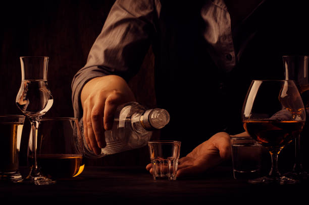 el camarero vierte el vodka o el tequila en una pequeña copa en el viejo mostrador del bar. vintage fondo de madera en pub o bar, estado de ánimo nocturno. lugar para texto, tonción, enfoque selectivo - tequila reposado fotografías e imágenes de stock